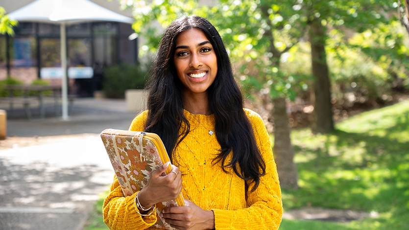 Female student in yellow jumper holding laptop case outside